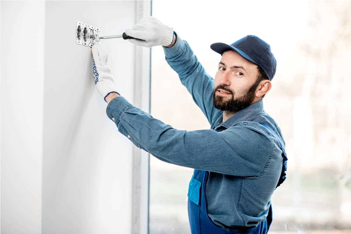 electrician in uniform mounting electric sockets on the white wall indoors